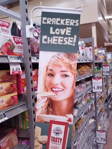 A supermarket advertisement depicting a very blonde white lady eating cheese and crackers, with the unfortunate slogan, "Crackers love cheese!" directly above her face.