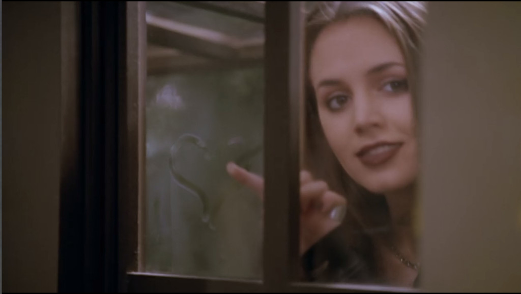 Faith is standing behind a window, having just drawn a heart on the glass. She's looking at Buffy with a very romantic look.