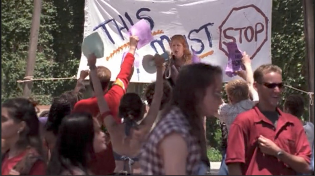A woman with a bullhorn stands in front of a bedsheet sign that reads "This must stop" while riled up students cheer her on.