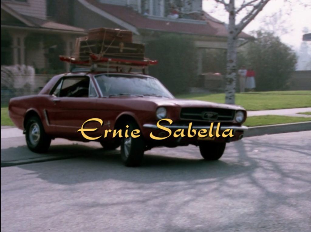 Larry's red Ford Mustang leaves the driveway. In the background, the out-of-focus figure of a woman in a red shirt stands beside a man. Another person in a red shirt is visible on a second-story balcony.