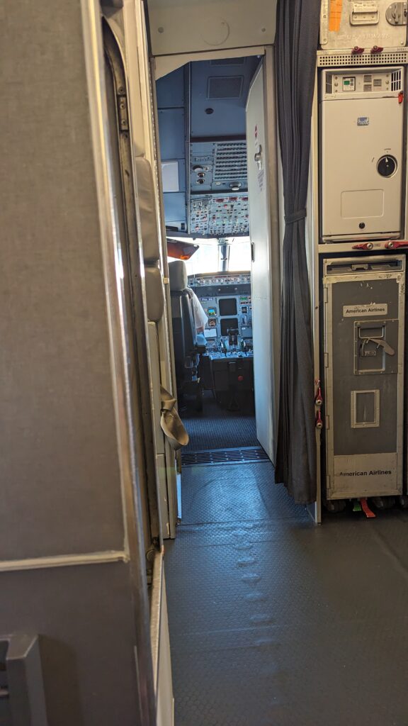 A view from the first row of a plane showing the aisle and the open cockpit door. Inside the cockpit are just like, so many buttons to push.