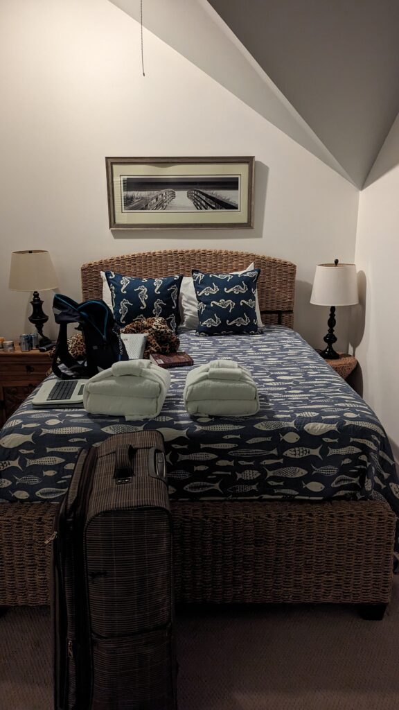A nice little finished attic room with a bed that has a lovely wicker headboard and and two bedside tables. All of my luggage is on everything. Above the bed is a photo of a boardwalk leading down to the beach.